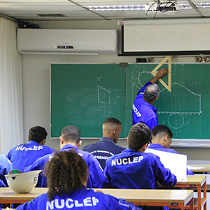 Imagem da galeria de imagem da página do centro de treinamento. A imagem mostra vários alunos sentados em suas cadeiras em uma sala de aula, tendo como principal assunto o treinamento e capacitação de pessoas.. O professor está ao fundo realizando cálculos geométricos em um quadro de giz.