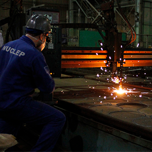 Imagem da galeria de imagem da página de Energia. A imagem mostra um funcionário de costas, manuseando um equipamento de corte para o corte de uma chapa metálica. O funcionário está de jaleco e equipamento de proteção.